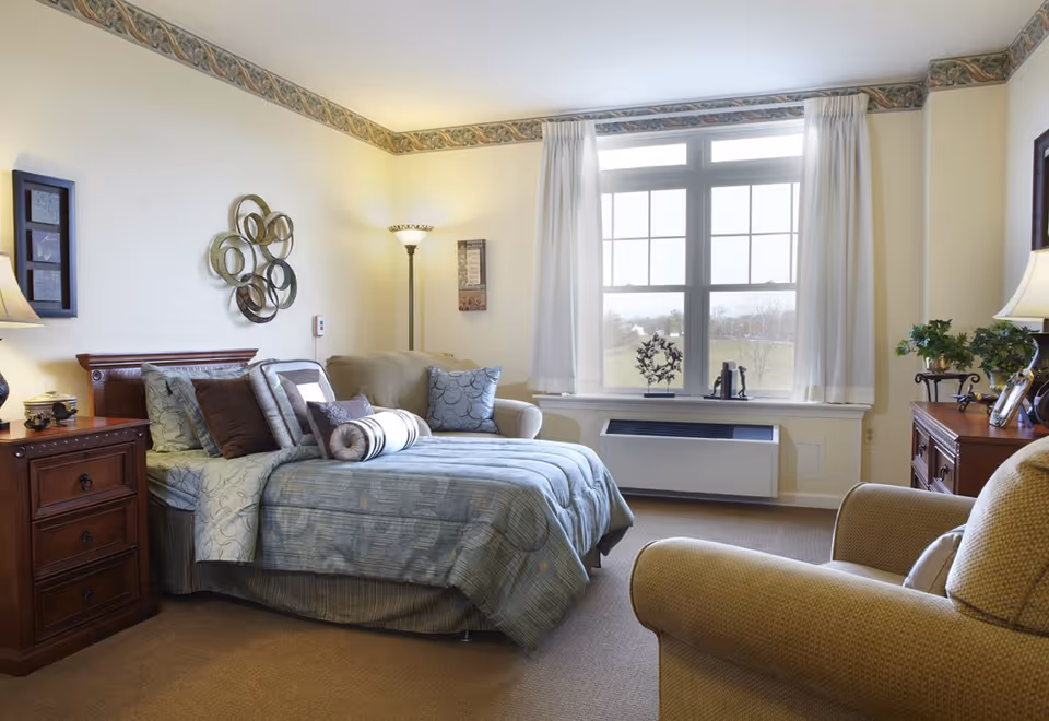 Sunny furnished bedroom with a made bed, armchairs, nightstands, and a large window overlooking a lawn.