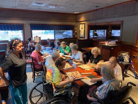 A group of elderly individuals seated around a long dining table in a well-lit room, engaging with menus or papers. A staff member stands nearby, and some residents use wheelchairs or walkers. The room has large windows with blue valances and wood-paneled walls.