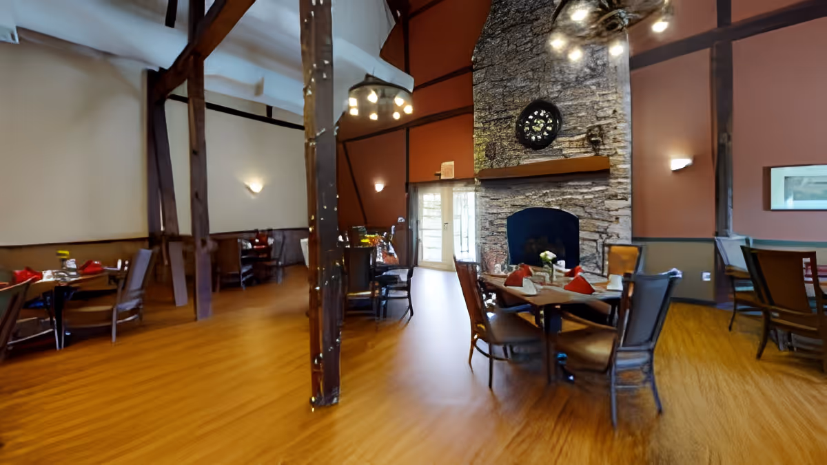 A dining room with wooden floors and exposed wooden beams. Several tables are set with chairs around them, some tables have red napkins and white table settings. A large stone fireplace with a circular decorative window above it is visible on one wall. The room has warm lighting and a cozy atmosphere.
