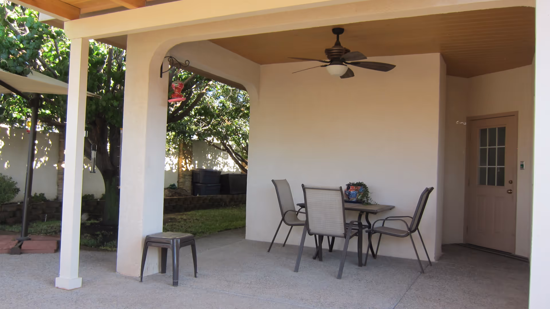 Covered outdoor patio area with a ceiling fan, a table with four chairs, a small stool, and a potted plant on the table. The patio overlooks a garden with trees and a white fence.