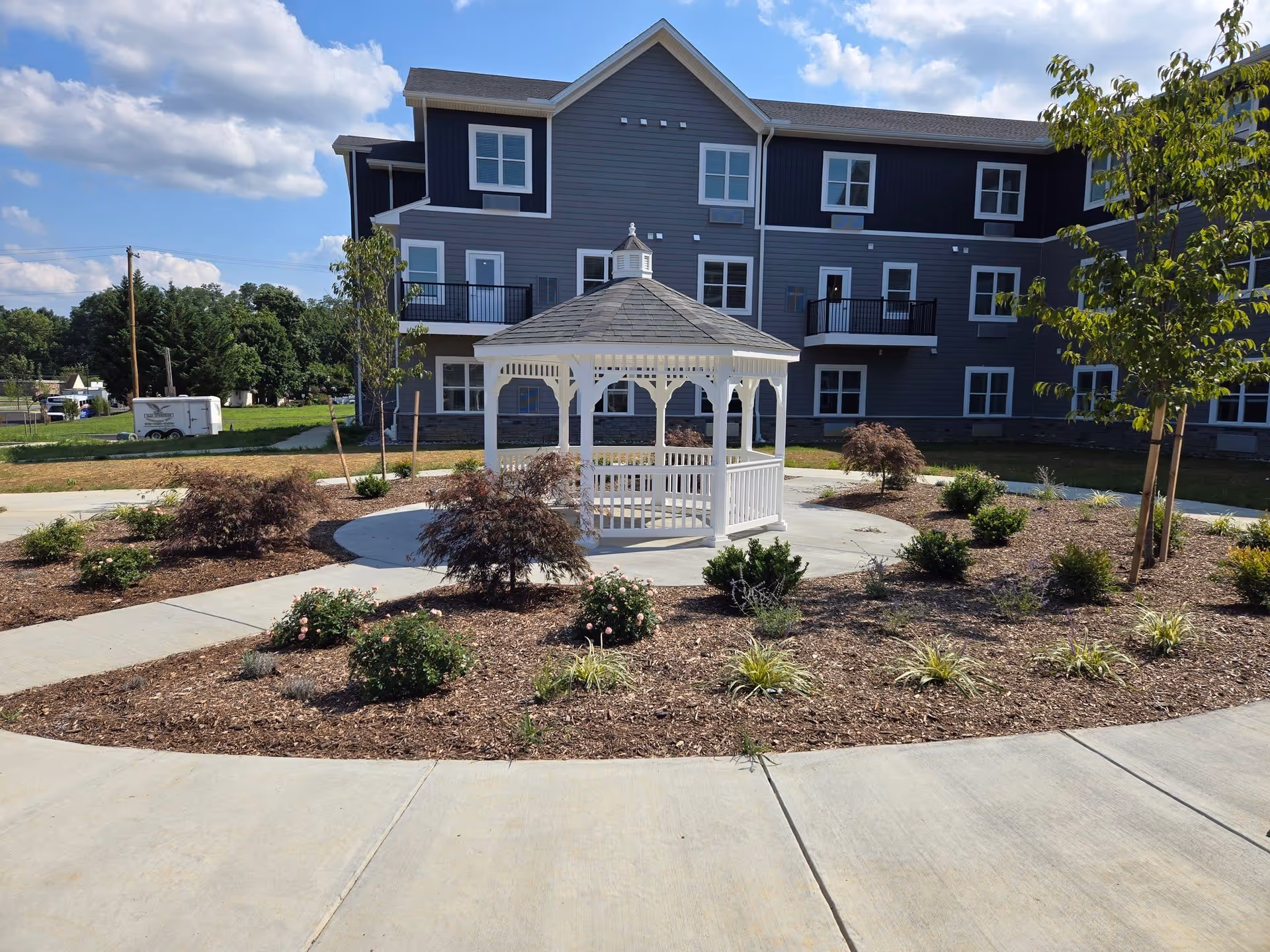 Outdoor garden area with a white gazebo in the center surrounded by landscaped plants and small trees, with a multi-story residential building in the background under a partly cloudy sky.