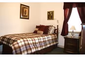 Small bedroom with a single bed dressed in plaid bedding, a nightstand with a lamp, framed art, and a curtained window.