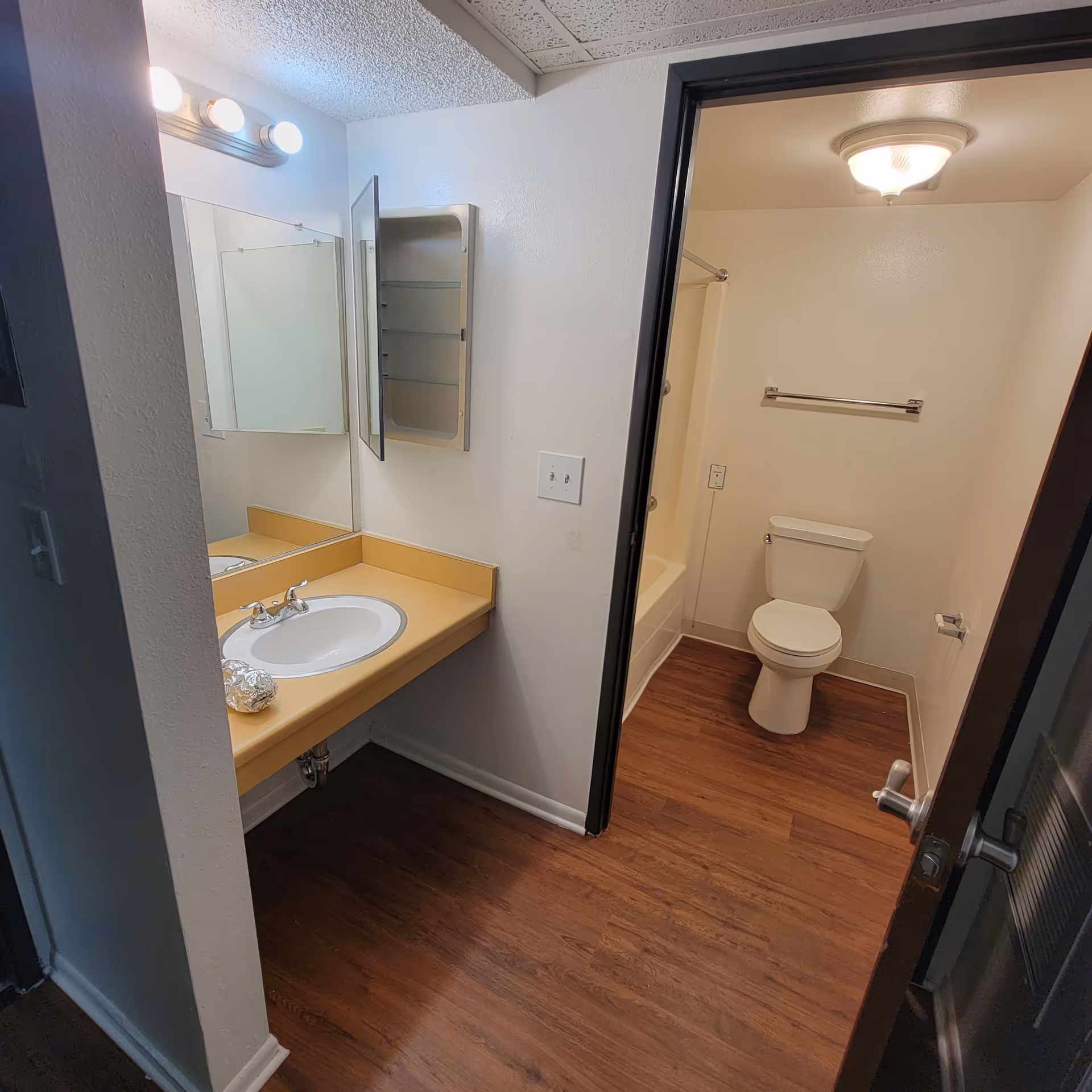 A bathroom with a wooden floor, a white toilet, a bathtub with a shower curtain rod, a sink with a yellow countertop, a mirror above the sink, and a wall-mounted medicine cabinet with its door open. The room is lit by ceiling and vanity lights.
