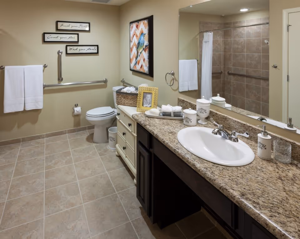 A clean and spacious bathroom with beige tiled floor and walls. It features a white toilet with safety grab bars around it, a large granite countertop with a white sink, soap dispenser, and decorative containers. A large mirror is mounted above the sink. On the wall above the toilet, there are three framed signs with hygiene reminders and a colorful bird painting. White towels hang on a towel rack and ring.