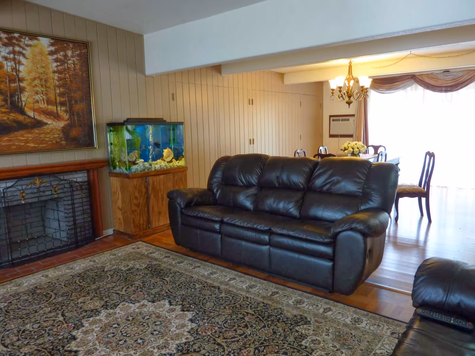 A living room featuring a black leather sofa, patterned area rug, fireplace with painting above, an aquarium, and a dining area beyond.