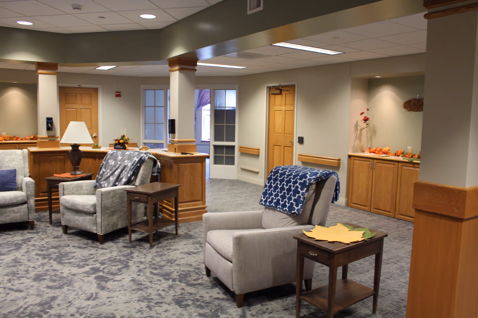 A cozy common area in a senior living facility with comfortable armchairs draped with blankets, wooden side tables, a lamp, and decorative autumn leaves on the tables. The room has beige walls, wooden trim, and multiple doors leading to other rooms.