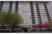 Exterior view of a multi-story residential building with numerous windows and balconies. In front of the building, there are trees with green and pink foliage, a small water fountain, and steps leading up to the entrance.