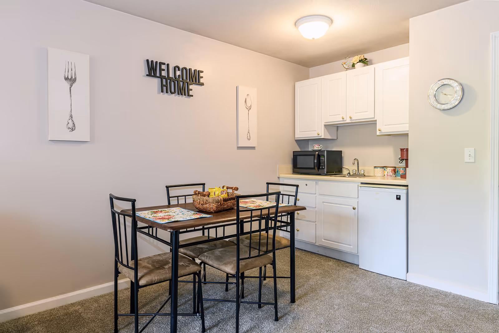 A small dining area with a wooden table and four metal chairs with cushioned seats. The table has two floral placemats and a basket with snacks. On the wall above the table, there are two framed pictures of a fork and a spoon, and a decorative sign that reads 'WELCOME HOME'. To the right, there is a kitchenette with white cabinets, a microwave, a small sink, a mini fridge, and a coffee maker. A round wall clock is mounted on the wall near the kitchenette.