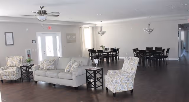 A spacious, well-lit common area with a light gray sofa and two patterned armchairs arranged around two small black side tables. In the background, there are two dining tables with chairs, a ceiling fan, and two chandeliers. The walls are light-colored, and there is a door with glass panels letting in natural light.