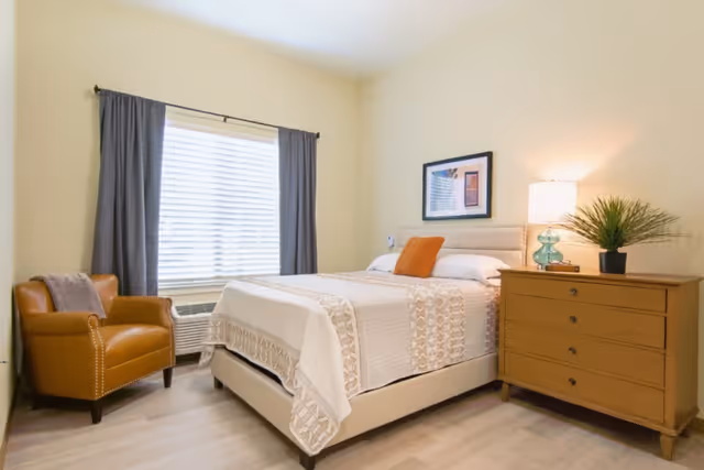 A bright and cozy bedroom featuring a neatly made bed with white and beige bedding, an orange accent pillow, a wooden dresser with a lamp and a potted plant, a framed picture on the wall, a window with gray curtains, and a brown leather armchair.