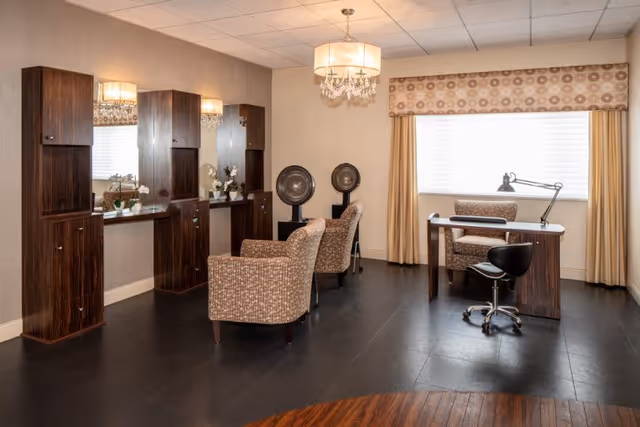 Interior view of a salon area in a nursing and rehabilitation center featuring two hair drying stations with chairs and mirrors, a desk with a lamp and chair near a window with curtains, and a chandelier hanging from the ceiling.