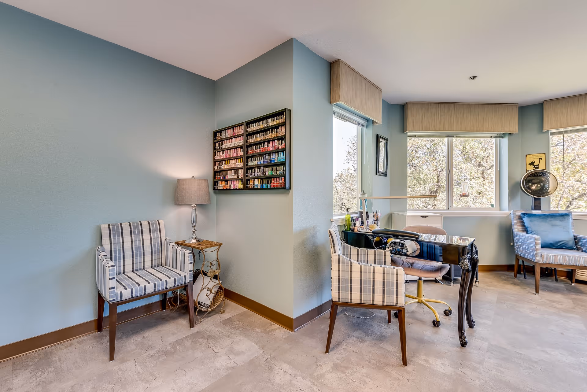 A bright room with light blue walls featuring a nail polish display mounted on the wall, a small side table with a lamp, and several chairs including a striped armchair and a salon chair at a black manicure table. Large windows with beige valances let in natural light and show trees outside.