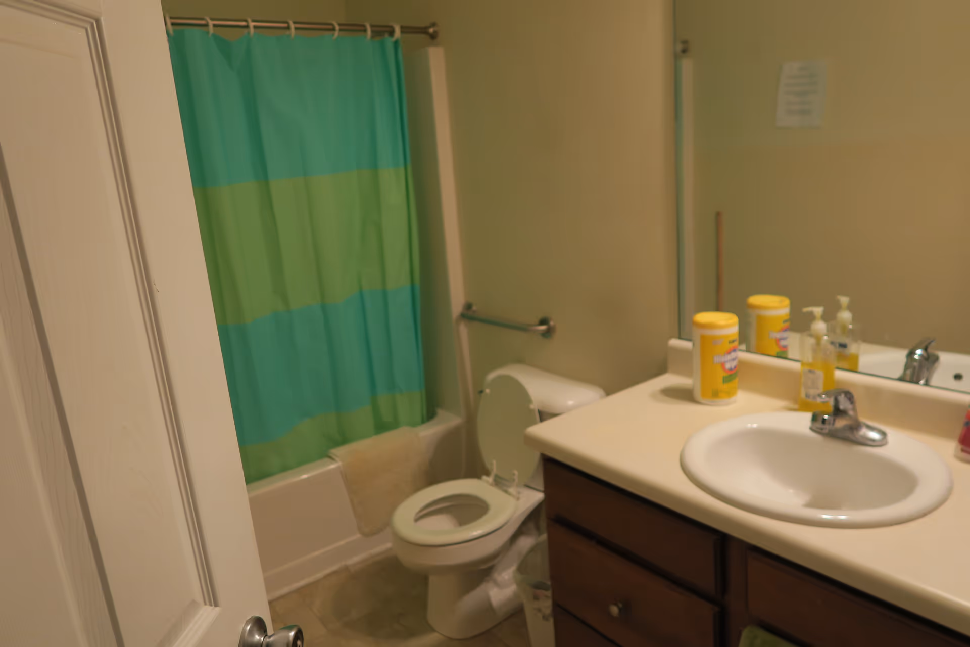 A bathroom with a white toilet, a bathtub with a green and blue striped shower curtain, a beige towel hanging on the bathtub, and a sink with a countertop holding disinfecting wipes and soap dispensers. A mirror is above the sink reflecting part of the room.