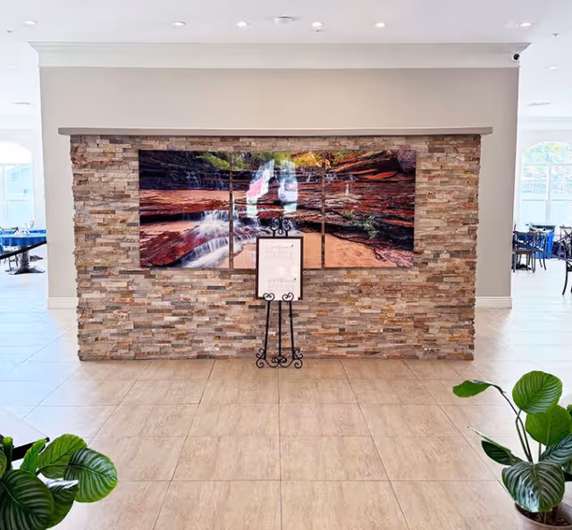 Stone feature wall displaying a large multi-panel waterfall painting above an easel sign in a tiled common area with plants.