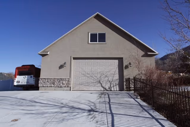 Front of a beige building with a closed two-car garage, a concrete driveway, a parked bus at left and a metal fence at right under a clear blue sky.