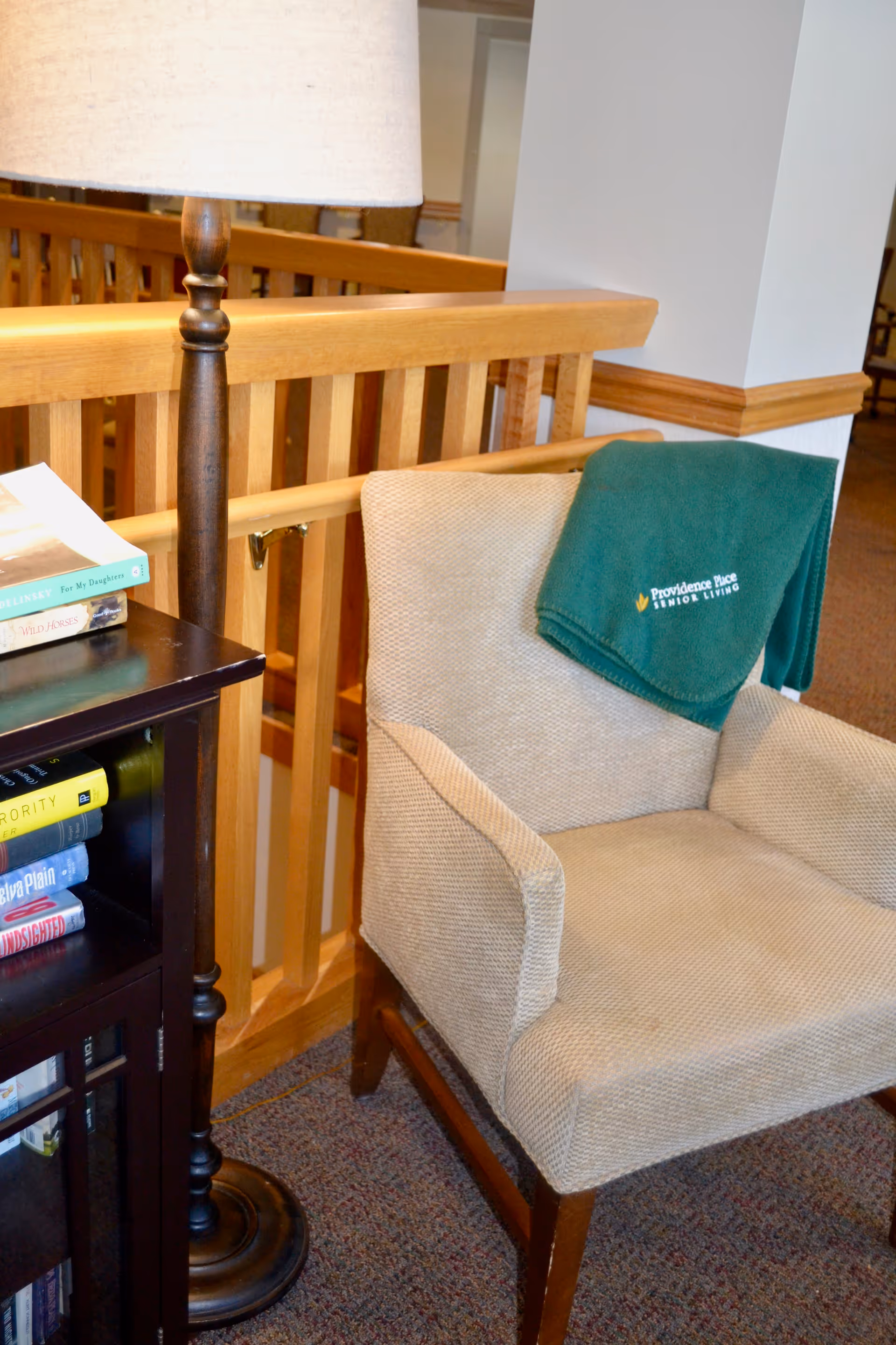Upholstered armchair with a green Providence Place Senior Living blanket draped over it beside a floor lamp, bookshelf, and wooden railing in a common area.