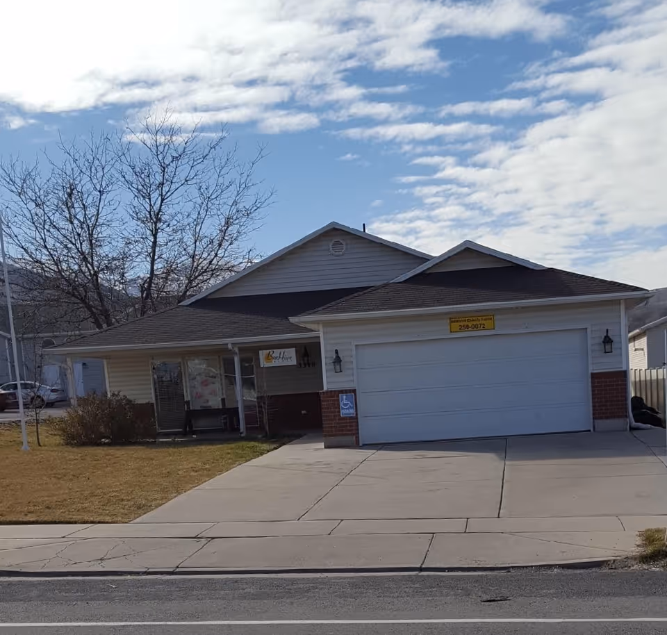 Single-story residential building with a two-car garage, front porch and driveway under a partly cloudy sky.