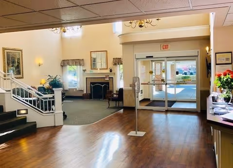 Interior view of a senior living facility lobby with wooden flooring, a staircase on the left, a seating area with chairs and a fireplace in the background, and glass automatic doors leading outside. There are framed pictures on the walls, a chandelier hanging from the ceiling, and a stand with hand sanitizer near the entrance.