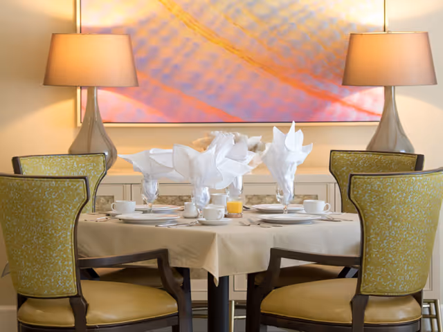 A dining table set for four with white napkins folded into decorative shapes, white plates, cups, and glasses, including a glass of orange juice. The table is covered with a beige tablecloth and surrounded by four green upholstered chairs with wooden frames. Two table lamps with beige shades are on a sideboard behind the table, and a large abstract painting with warm colors hangs on the wall above the sideboard.