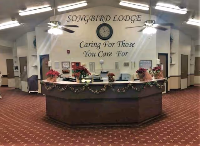 Reception desk area inside Songbird Lodge with a curved counter decorated with garlands and poinsettia plants. Behind the desk is a wall with a clock and the text 'SONGBIRD LODGE Caring For Those You Care For'. The area has a patterned carpet, ceiling fans with lights, and several doorways leading to other rooms.