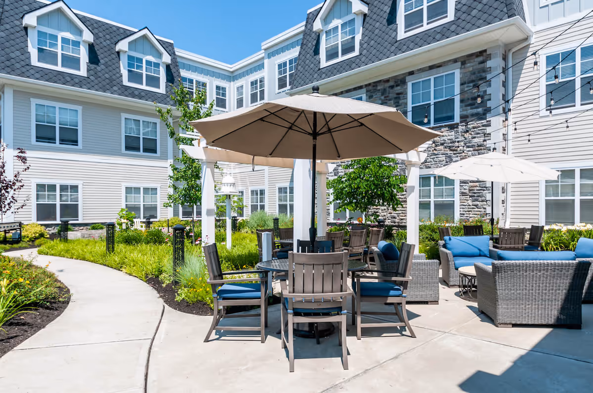 Outdoor patio area at Arbor Terrace Mountainside featuring a round table with wooden chairs and blue cushions under a large beige umbrella. Surrounding the patio are additional seating areas with wicker furniture and blue cushions, string lights overhead, and a well-maintained garden with green plants and flowers. The background shows a multi-story building with gray and beige siding, stone accents, and multiple windows under a clear blue sky.