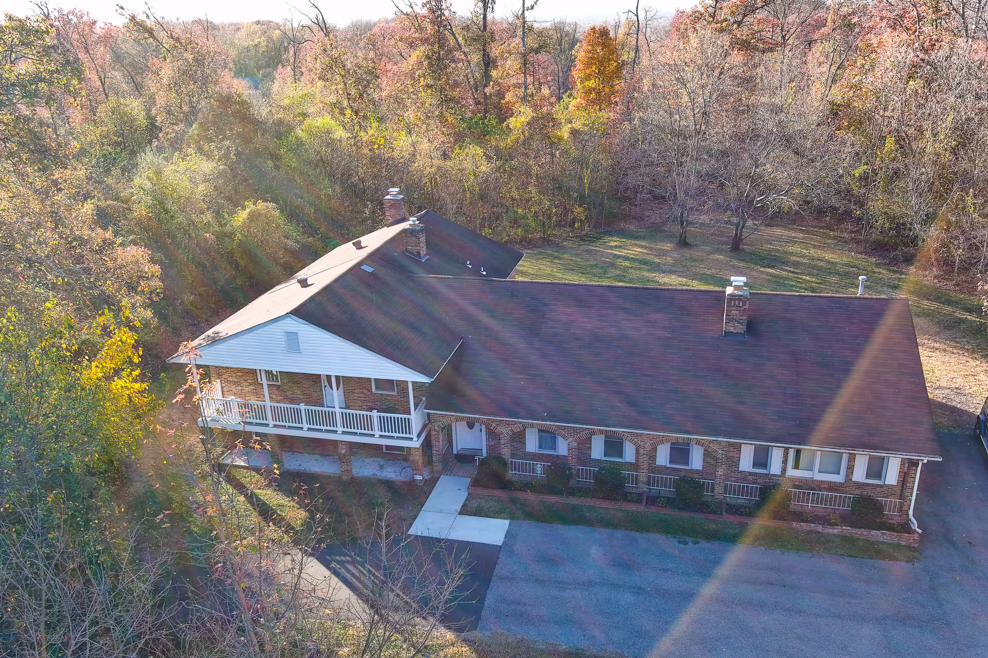 Aerial view of a single-story brick building with a dark roof surrounded by trees with autumn foliage. The building has a small porch with white railings and a paved driveway in front.