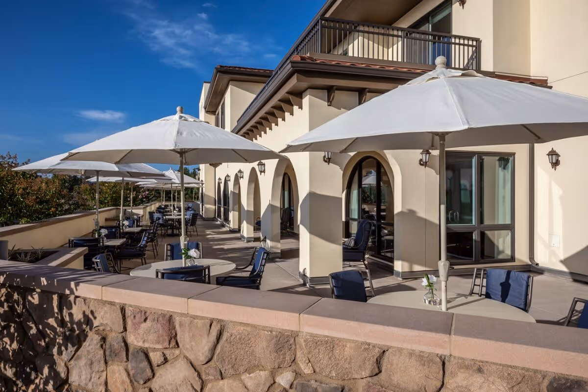 Outdoor patio area at Westmont of Carmel Valley with multiple round tables and chairs under large white umbrellas, adjacent to a beige building with arched doorways and windows, under a clear blue sky.