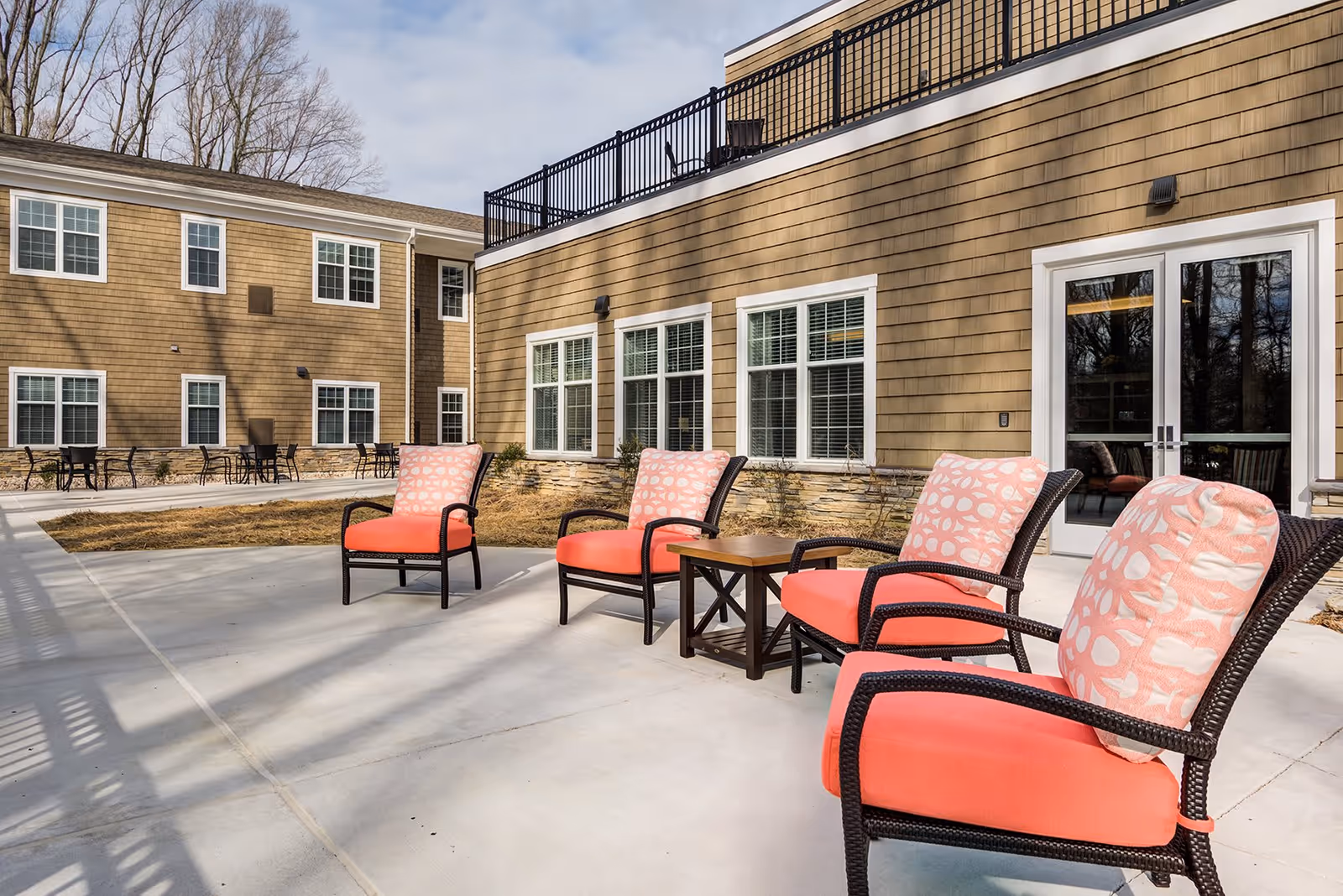 Outdoor patio area at a senior living facility with four cushioned chairs featuring coral-colored seats and patterned back cushions arranged around a small wooden table. The building has tan siding with white-framed windows and a glass door leading inside. Leafless trees are visible in the background under a partly cloudy sky.