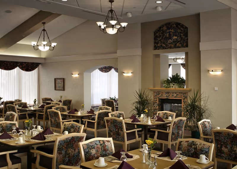 A dining room with multiple tables set with cups, glasses, and folded napkins. The chairs have floral upholstery. The room features a fireplace with plants on either side, wall sconces, chandeliers, and large windows with curtains allowing natural light.