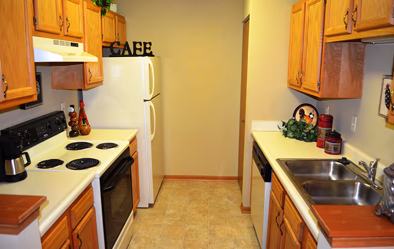 A narrow kitchen with wooden cabinets on both sides, a white electric stove and oven on the left, a white refrigerator at the end, and a double stainless steel sink on the right. The countertops are light-colored, and there are decorative items including a sign that says 'CAFE' on top of the refrigerator and some small plants and containers on the counters.