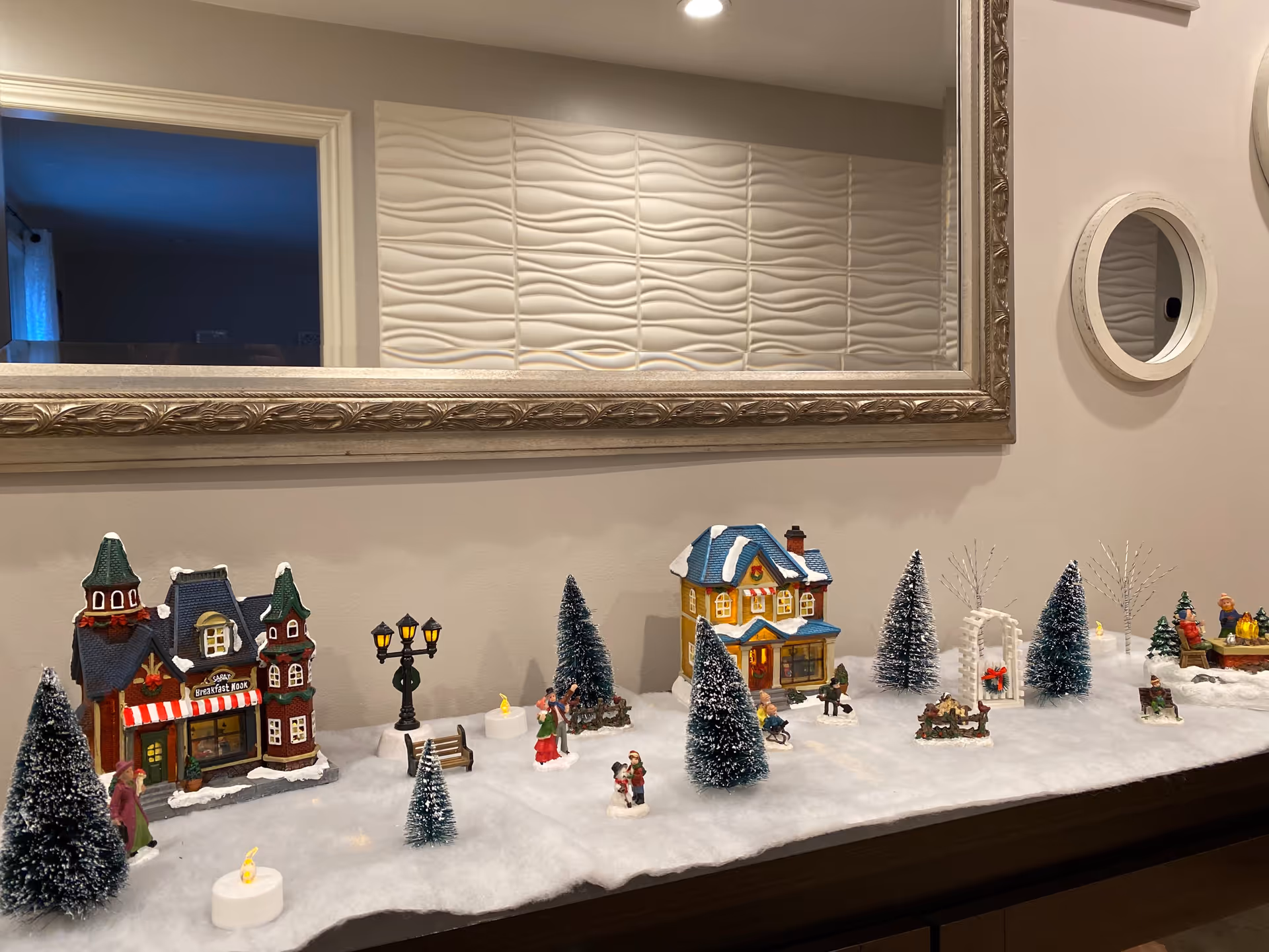 A decorative winter village scene displayed on a wooden surface with miniature houses, snow-covered trees, small figurines of people, benches, street lamps, and artificial snow. The background features a large ornate mirror and a textured wall panel.