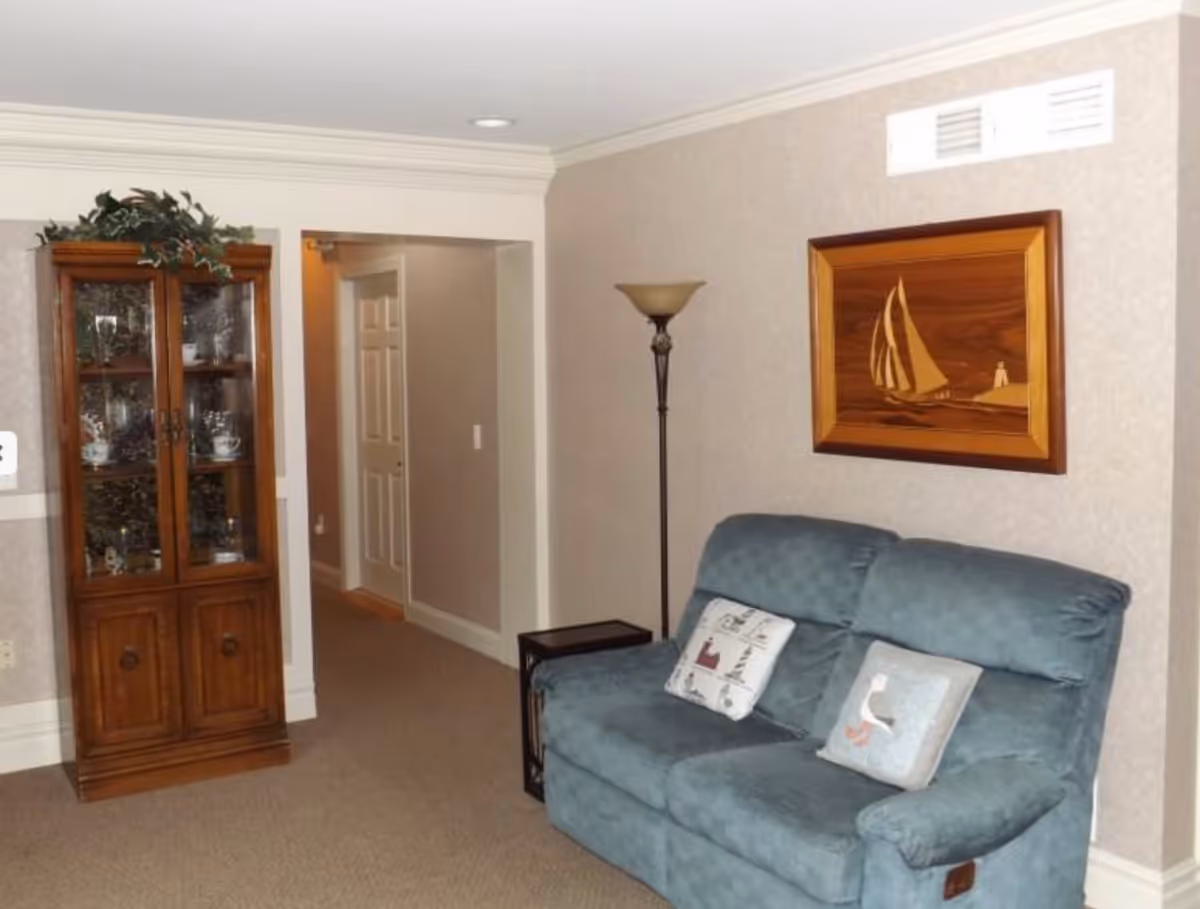 A cozy living room area with a blue upholstered loveseat featuring two decorative pillows. To the left, there is a wooden cabinet with glass doors displaying china and decorative items. A tall floor lamp stands next to the loveseat, and a wooden framed artwork of a sailboat hangs on the wall above it. The room has beige walls and carpeted flooring, with a hallway visible in the background.