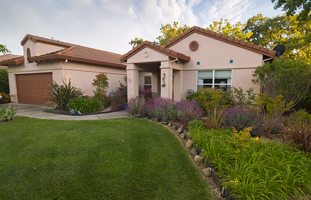 Single-story house with a tiled roof and a two-car garage, surrounded by a well-maintained garden with green grass, various shrubs, and flowering plants under a partly cloudy sky.