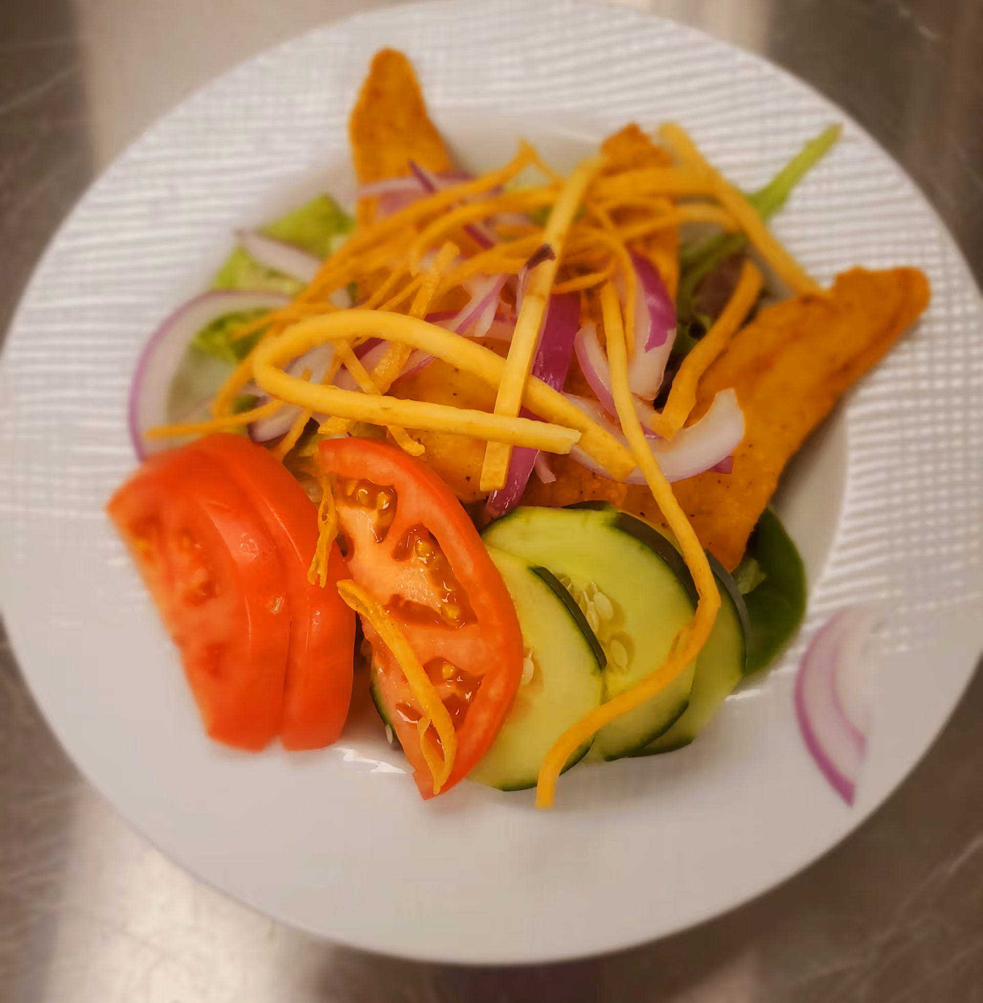 A white plate with a salad consisting of sliced tomatoes, cucumbers, red onions, crispy fried strips, and pieces of seasoned fried chicken on a bed of lettuce.