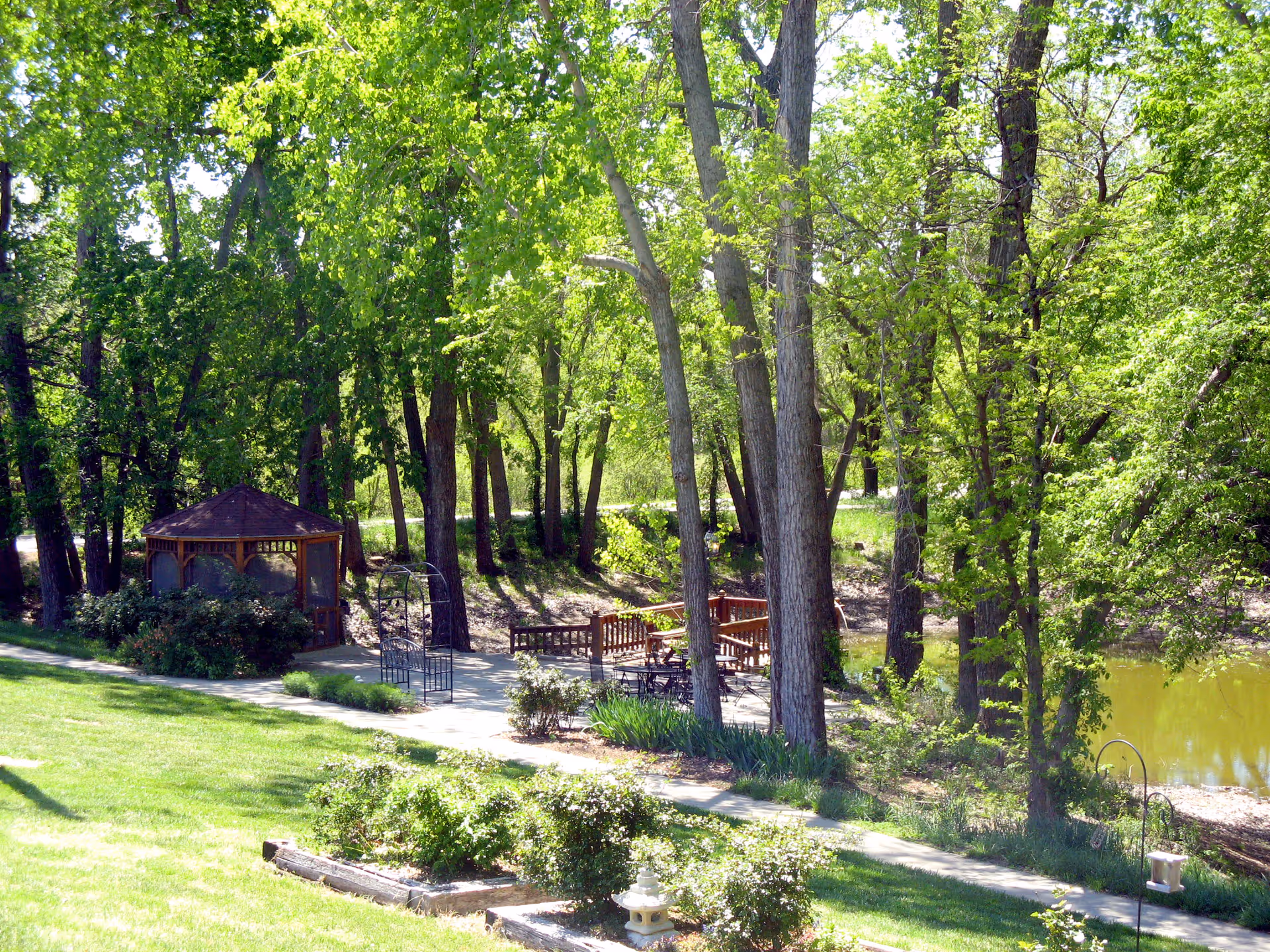 A shaded garden area with a wooden gazebo, benches and a pathway beside trees and a small pond.