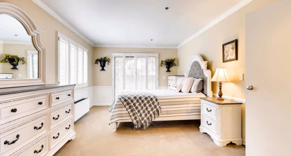 A bright and spacious bedroom with a large bed featuring a tufted headboard, striped bedding, and a checkered throw blanket. The room has beige walls, carpeted floor, a dresser with a large mirror, two nightstands with lamps, and windows with white plantation shutters allowing natural light to fill the space. There are decorative plants mounted on the walls and a framed picture above one nightstand.