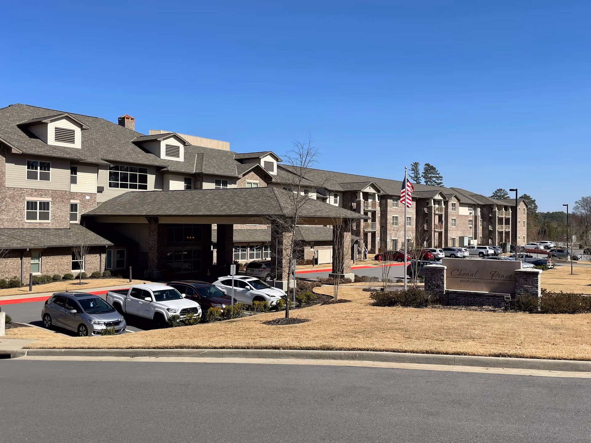 Exterior view of Chenal Pines Retirement Resort, a large multi-story building with a covered entrance and several parked cars in front. The building has a combination of brick and siding with multiple windows and dormers on the roof. An American flag is flying near the entrance, and a sign with the facility name is visible on the lawn.
