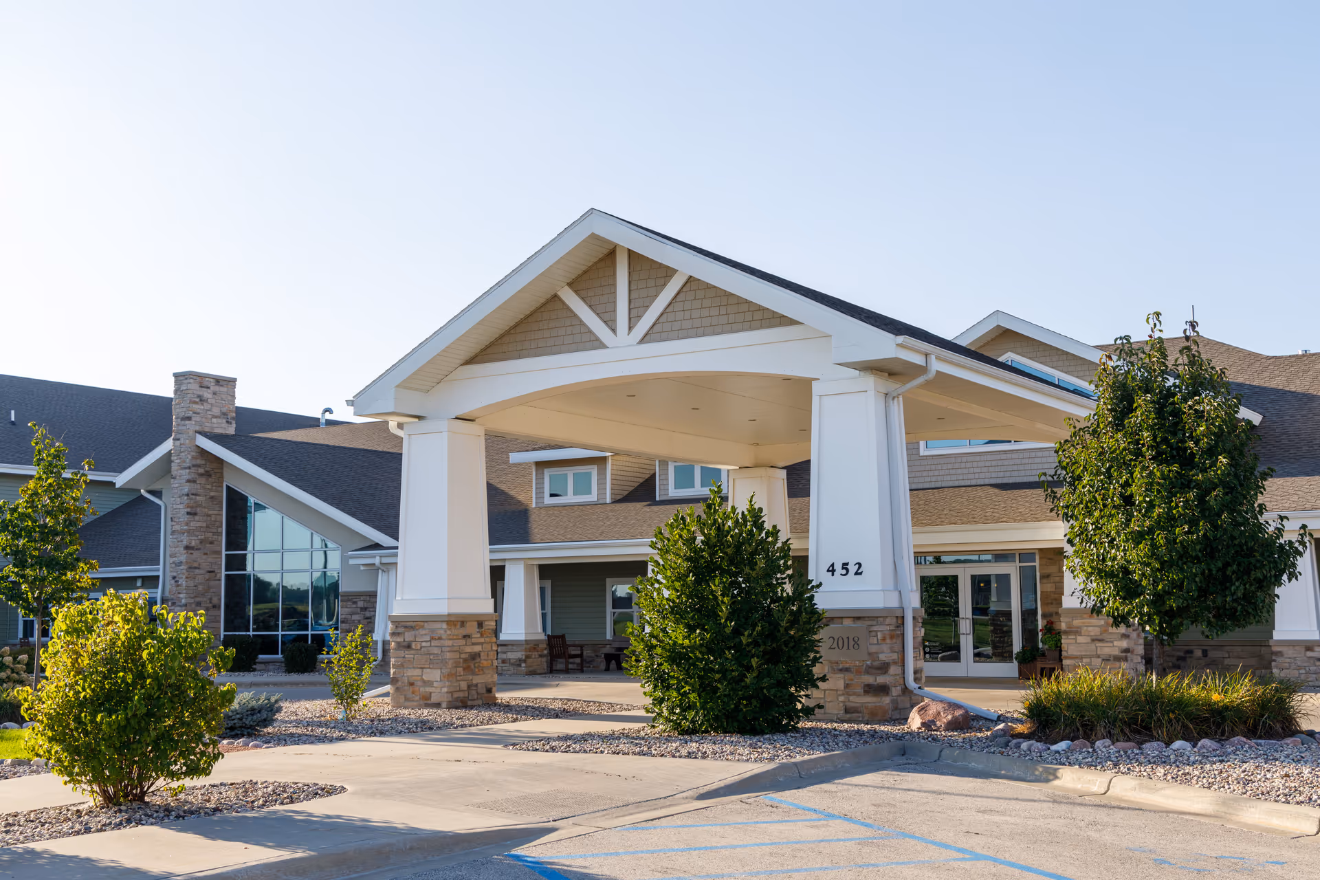Exterior view of the Christian Living Center building entrance with a covered drop-off area supported by large white columns with stone bases. The building has a pitched roof, large windows, and landscaping with bushes and trees around the entrance. The sky is clear and the parking area is visible in the foreground.