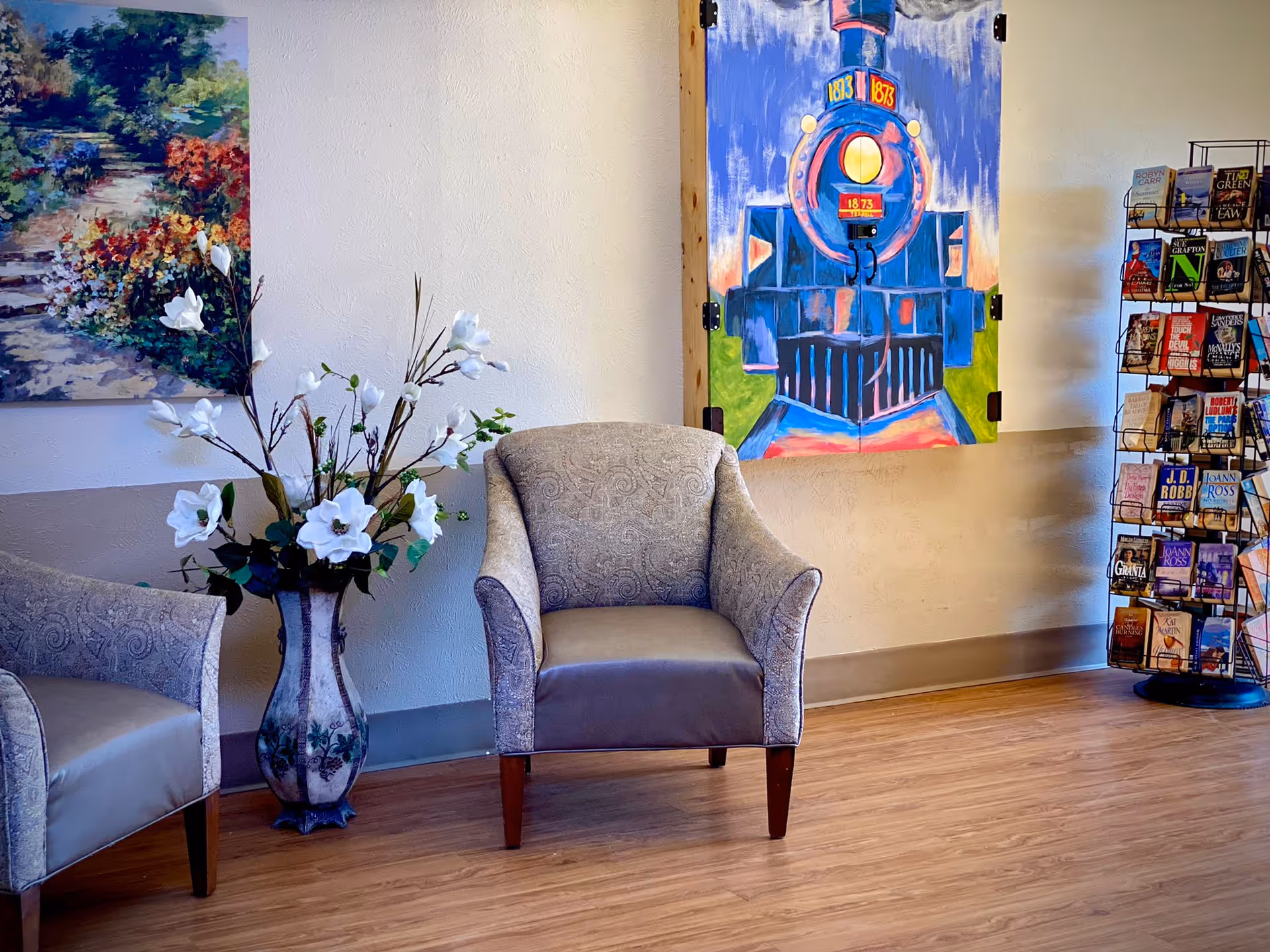 A cozy seating area with two upholstered armchairs, a tall vase with white flowers, a colorful painting of a train on the wall, and a rotating rack filled with various books in the corner.