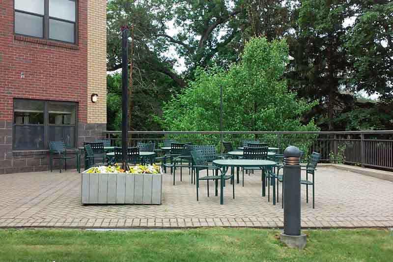 Outdoor patio area with several green metal tables and chairs arranged on a paved surface next to a brick building. There is a large planter with flowers and a tall pole in the middle of the patio. Trees and greenery are visible in the background.