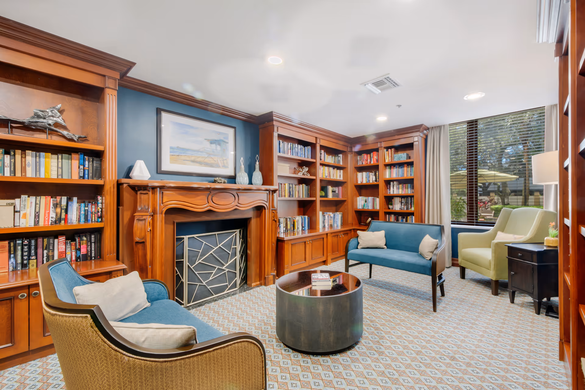 A cozy library room with wooden bookshelves filled with books, a wooden fireplace with a decorative screen, two blue cushioned sofas, a green armchair, a round coffee table, and a large window with blinds showing an outdoor patio area.