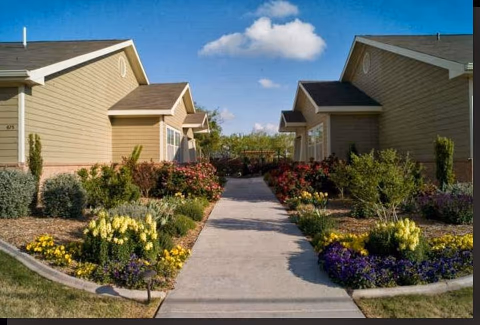 A paved walkway between two beige single-story buildings with gabled roofs, bordered by well-maintained flower beds featuring various colorful flowers and shrubs under a blue sky with a few clouds.