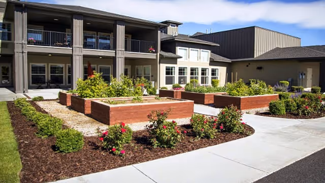 Outdoor garden area with raised wooden planter boxes filled with plants and flowers, surrounded by mulched flower beds and a concrete walkway. In the background, there is a two-story building with balconies and large windows under a blue sky with some clouds.