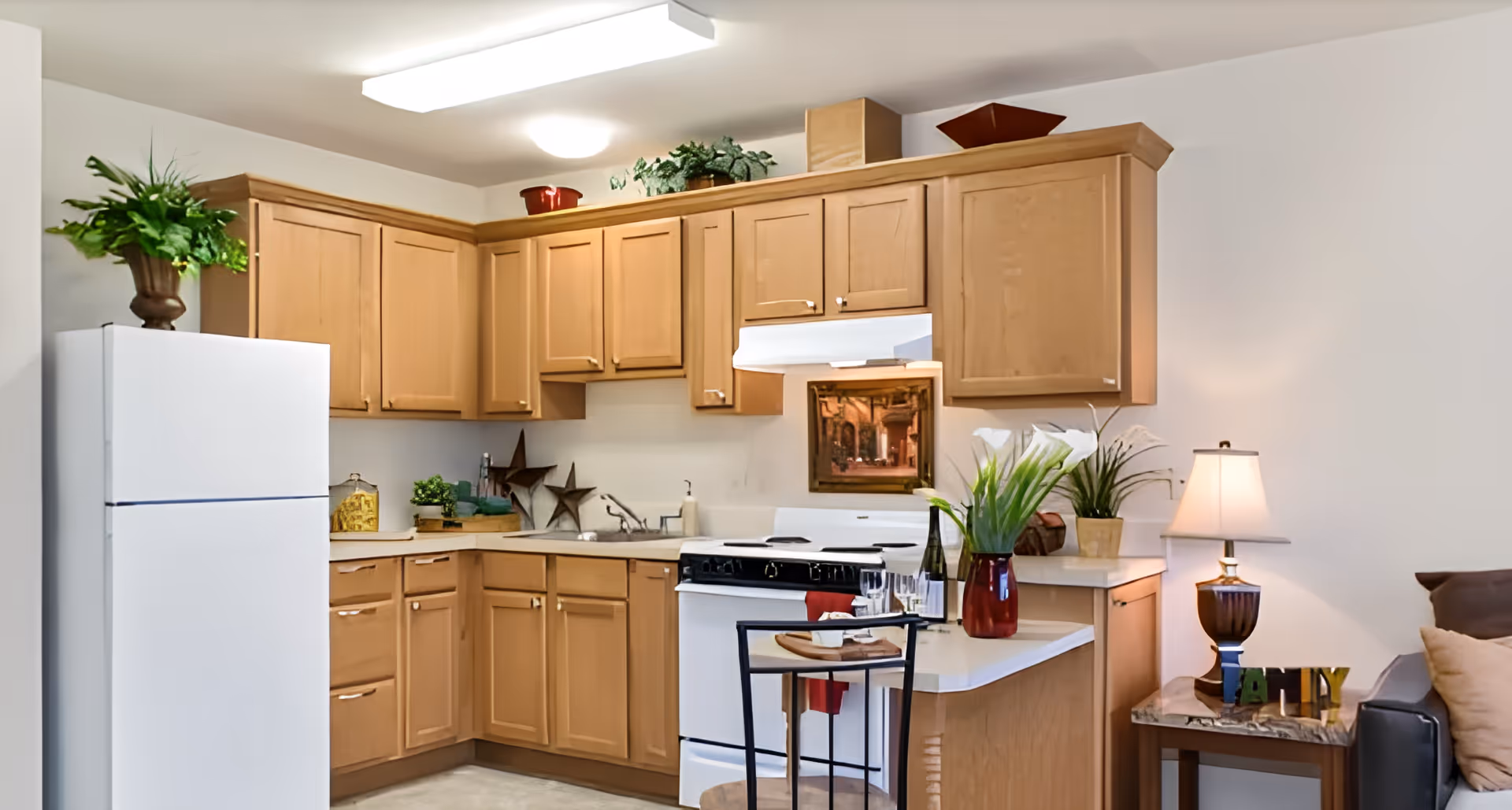 Open kitchen with light wood cabinets, a white refrigerator and stove, countertop seating, and decorative plants and lamps.