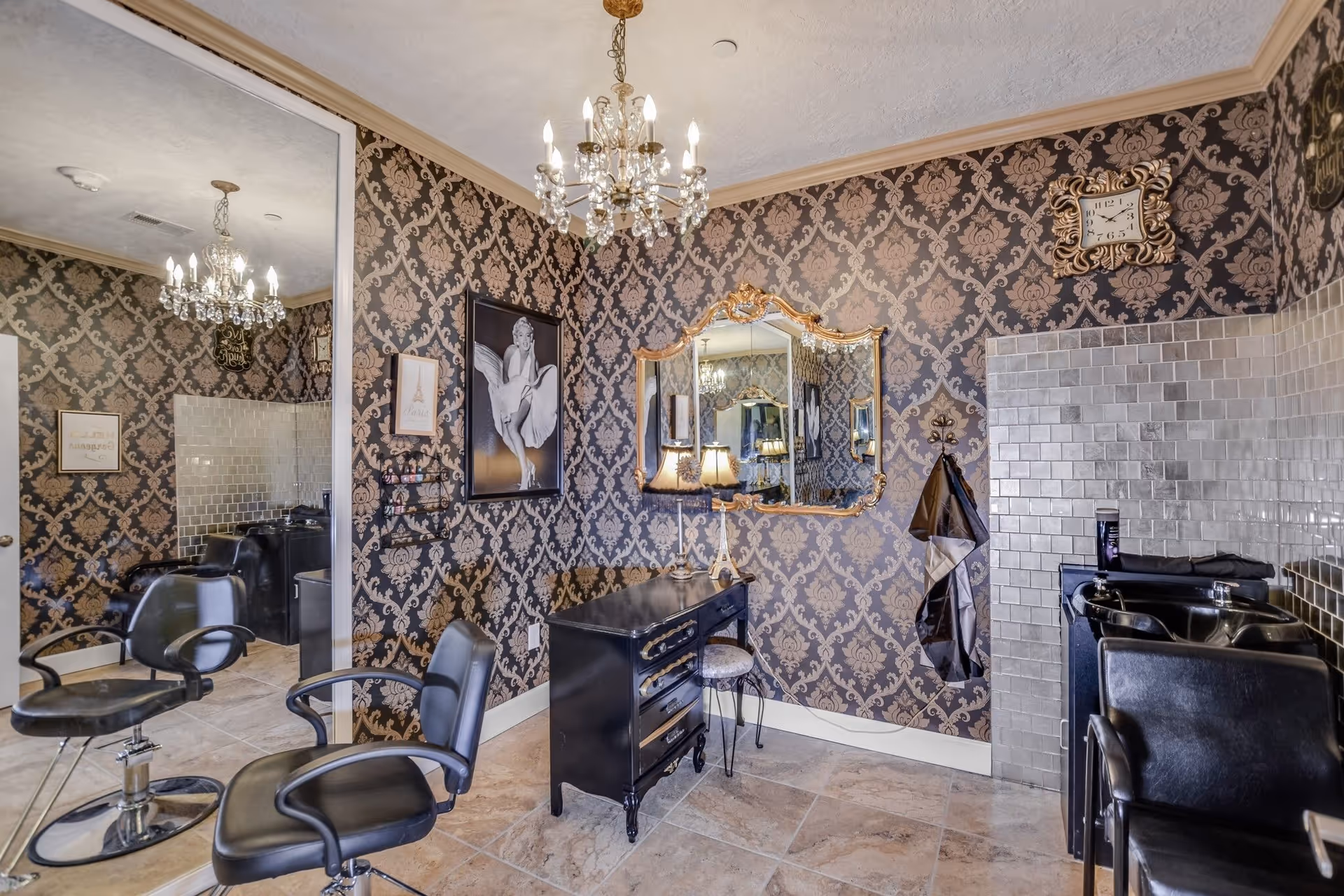 Interior of a stylish hair salon with black salon chairs, a black vanity with a stool, a large ornate mirror, and a chandelier hanging from the ceiling. The walls are decorated with patterned wallpaper and framed pictures, including a black and white photo of Marilyn Monroe. There is a tiled area with hair washing sinks and a clock on the wall.