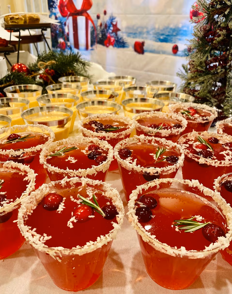 Close-up view of festive holiday drinks in clear cups with rims coated in shredded coconut, garnished with cranberries and rosemary sprigs, arranged on a table with additional yellow dessert cups and holiday decorations in the background.
