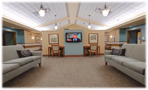 A spacious senior living common area with two gray sofas facing each other, each with a decorative pillow. The room has a high ceiling with multiple hanging light fixtures. At the far end, a flat-screen TV is mounted on a partition wall, flanked by two chairs and framed artwork on the walls. The space has a warm and inviting atmosphere with neutral tones and carpeted flooring.