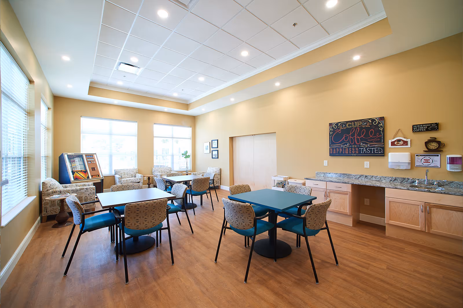 A bright and spacious common dining area with several tables and chairs arranged on a wooden floor. The room has large windows with blinds allowing natural light to fill the space. There is a countertop with a sink and cabinets along one wall, decorated with coffee-themed signs. Comfortable armchairs are placed near the windows, and a vintage jukebox is visible in the corner.