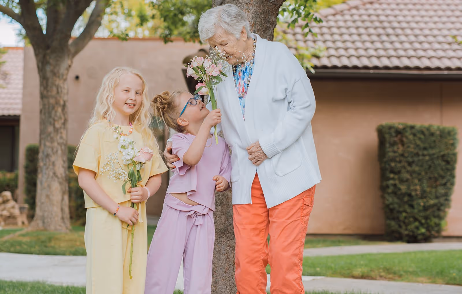 An elderly woman standing outdoors with two young girls, all holding flowers. They are near a tree with a building in the background, and the scene appears to be in a garden or courtyard area.