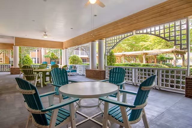 Covered outdoor seating area with round tables and green Adirondack chairs, overlooking a garden with trees and pergolas. The space has a ceiling fan and decorative wooden latticework along the railing.